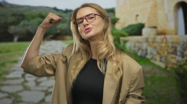 Gözlüklü, güçlü kollu bir kadın, açık havada, güneşli bir parkta bej ceketli ve siyah gömlekli, bulanık doğal bir arka plana karşı mutluluk ve güç gösteren bir jest yapıyor..