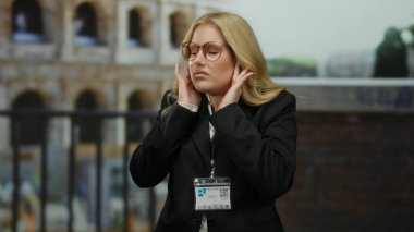 Gözlüklü bir kadın Roma 'daki stadyumdan iğreniyor siyah bir takım elbise ve kimlik kartı takıyor, sarı saçlı, tarihsel bir arkaplana karşı beklenmedik bir tepki gösteriyor..