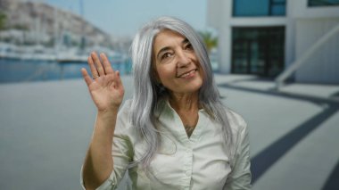Gri saçlı yaşlı kadın, güneşli gökyüzünün altındaki deniz kenarında mutlu bir şekilde el sallıyor. İçtenlik ve dostluk gösterisi yapıyor..