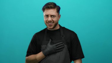 Önlük ve eldiven giyen genç sakallı beyaz adam karizmatik ve etkileyici bir şekilde yeşil duvara karşı poz veriyor. Muhtemelen bir garsonu ya da aşçıyı temsil ediyor..