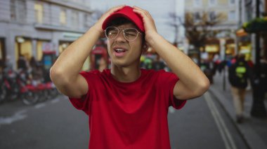 Kırmızı kurye üniforması giyerken trafiği bulanıklaştıran kalabalık bir sokak sahnesinde iki eliyle başını tutuyor..