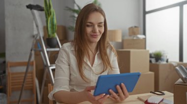 Kadın, binadaki karton kutular ve merdiven arasında gülümserken tablet tutuyor; heyecan başarısı.