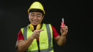 Kasklı ve güvenlik yeleği olan gülümseyen inşaatçı elinde siyah arka plan aydınlatma cihazıyla ürünü vurguluyor.