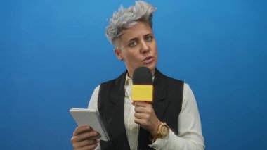 Kısa gri saçlı, notlar okuyan ve mikrofonu parlak mavi stüdyoda tutan bir kadın..