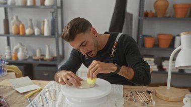 Kapalı mekandaki zanaatkar atölyesinde elleri dönen bir çanak çömlek üzerinde kili şekillendiren adam; dikkatli yaratıcılık.