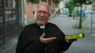 Siyah cüppeli kıdemli rahip elinde İncil ile sokak ortasında neşeli bir ifadeyle ve gündüz vakti şehir ortamında davetkar bir jest yapıyor.