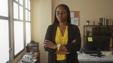 Sarı tişörtlü ve siyah gözlüklü kadın, ofis ekipmanları ve geniş pencereli modern ofis ortamında, kendinden emin bir şekilde duruyor..