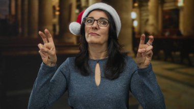 Orta yaşlı İspanyol kadın kilisenin içinde iki eliyle kırmızı Noel Baba şapkası takarak tırnak işareti yapıyor..