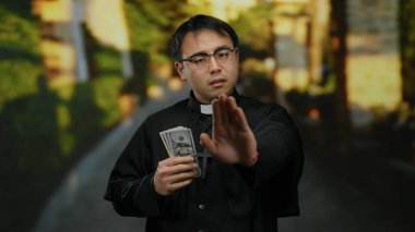 Asyalı rahip elinde para, gözlük ve siyah bornoz takıyor, el hareketleriyle inkar ediyor, dini ve finans konularını temsil ediyor..