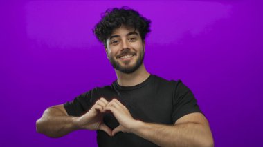 Sakallı genç İspanyol adam kalp hareketi yapıyor. Mor arka plana gülümsüyor. Sevgi ve pozitifliği ifade ediyor.