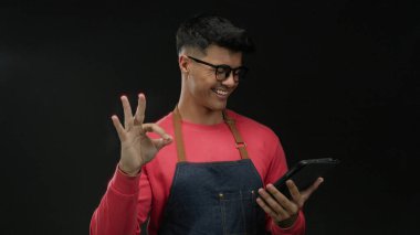 Gözlüklü ve önlüklü genç adam karanlık arka plan karşısında tabletle etkileşim içinde modern teknolojiyi ve moda tarzını sergiliyor güzellik ve basitliği vurguluyor.