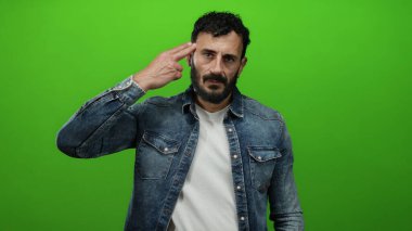 Sakallı İspanyol bir adam yeşil arka planda jest yapıyor, kot ceket ve beyaz gömlek giyiyor..