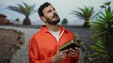 Turuncu tulumlu İspanyol adam yeşil ağaçlarla çevrili açık hava parkında İncil 'i tutuyor ve doğada dururken inancı yansıtıyor..