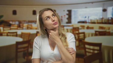 Parlak bir restoranda, boş masa ve sandalyelerle çevrili, yüzünde merak ve düşüncenin ifadesi olan bir kadın.