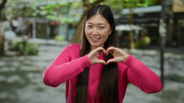 Pembe kazaklı bir kadın şehir sokaklarında elleri ile kalp şekli yapıyor. Neşenin, mutluluğun ve aşkın vücut bulmuş halidir..