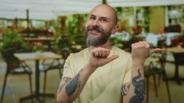 Sakallı ve dövmeli bir adam şehir restoranındaki terasta neşeli bir şekilde el kol hareketi yapıyor.