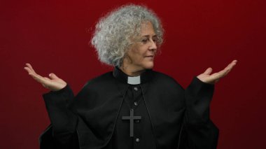 Gri saçlı, rahip kıyafeti giyen, kırmızı arka planda el ele tutuşan kadın sakin ve otoriter görünüyor.