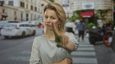 Sarışın kadın şehir merkezinde yoğun bir caddede sert bir ifadeyle kamerayı işaret ediyor. Cesur bir meydan okuma..