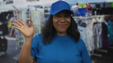 Mavi üniformalı kadın kurye, hediyelik eşya tezgahları ve giysi raflarıyla dolu açık hava pazarında el ele sallıyor. Dostluk..