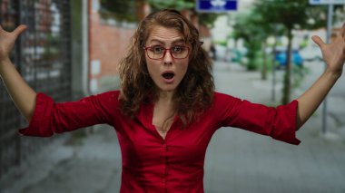 Kırmızı gömlekli kadın kıvırcık saçlı ve gözlüklü duygularını ifade ediyor. Gündüz vakti açık havada sürpriz olduğunu gösteriyor..