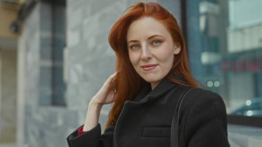 Şehirli bir caddede, uzun kızıl saçlı ve siyah ceketli bir kadın güvenle gülümsüyor. Şehir zeminine karşı. Modern bir yaşam tarzıyla güzellik ve moda sergiliyor..