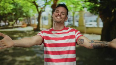 Parkta sırıtan bir adam çizgili bir tişört giymiş, arka planda ağaçlarla sevinçle birbirine kenetlenmiş mutluluk ve memnuniyeti ifade eden bir adam..