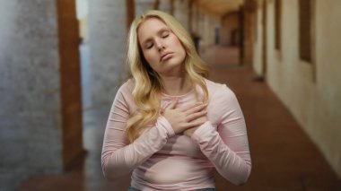 Üniversite koridorunda elleri göğsünde duygularını ifade eden bir kadın. Eski bir kampüs arka planında sarı saçlar ve düşünceli bir ifade sergiliyor..