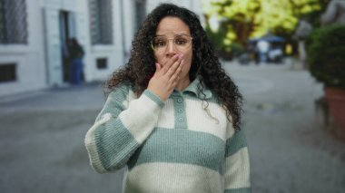 Gözlüklü bir kadın şehir manzarası ile çevrili bir sokakta şaşkınlığını ifade ediyor. Kıvırcık saçlar, duygular, günlük tarz, gündüz ifadesi ve gençlik ifadeleri sergiliyor..