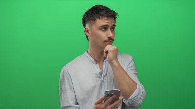 Genç adam canlı yeşil arka plan önünde akıllı telefonu tutarken, modern teknolojiyi ve içsel düşünceleri tasvir ederken, gündelik açık gömlekler giyerken,.
