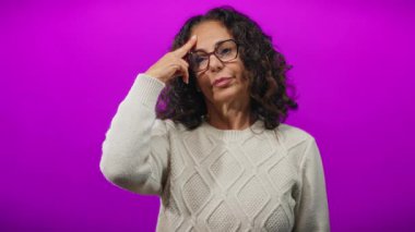 Kıvırcık saçlı ve gözlüklü İspanyol bir kadın parlak mor bir arka plana dokunuyor, düşünceli ve düşünceli bir ifade sergiliyor..