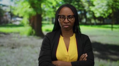 Gözlüklü ve kıvırcık saçlı bir kadın güneşli bir park ortamında hoşnutsuz görünüyor, sarı bir gömlek ve siyah bir ceket giyiyor, açık havada kollarını kavuşturmuş duruyor..