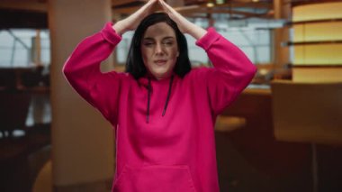 Kapalı mekanlarda gülümseyen pembe kapüşonlu bir kadın, ellerle üçgen yapıyor, esmer saçlı, modern bar ortamında günlük tarz, parlak ve hayat dolu bir ortam..
