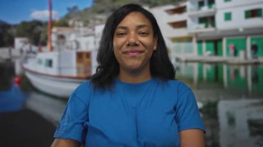 Parlak mavi tişörtlü bir kadın, demir atmış bir tekne ve sakin bir rıhtımın yanındaki bir limanda başparmağını işaret ediyor..