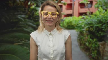 Parlak sarı gözlüklerle ayakta duran ve gülümseyen bir kadın yemyeşil çalıların yanında ve dolambaçlı bir yolda canlı bir parkta güven;.