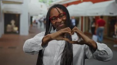 Gözlüklü kadın şehir sokağında kalp hareketi yapıyor. Gündüz vakti geçmişi bulanık.