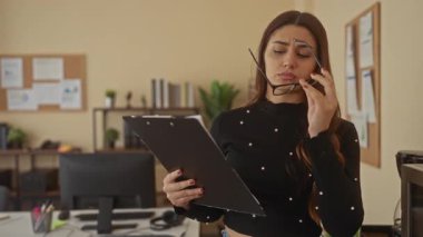 Modern ofis ortamında arka planda bitki ve kitaplarla dolu dokümanları incelerken elinde pano tutan kadın.