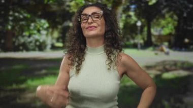 Kıvırcık saçlı genç İspanyol kadın ellerle kalp şekli oluşturan gözlük takıyor ve açık havada yeşil ormanda gülümsüyor..