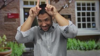 Çizgili gömlekli genç İspanyol adam gülümsüyor ve işaret parmaklarıyla binaya işaret ediyor..