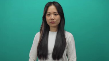 Uzun siyah saçlı bir kadın, yeşil bir arka plana karşı duruyor, beyaz bir gömlek giyiyor, kollarını kavuşturmuş, kararlı ve dengeli bir ifade sergiliyor..