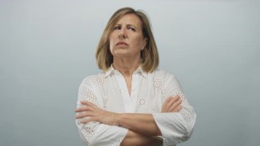 Stüdyoda soluk gri arkaplan karşısında çapraz kollu beyaz bluzlu, orta yaşlı beyaz bir kadın; şüphecilik.