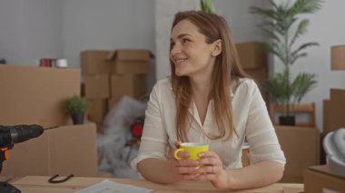 Beyaz kadın, elinde karton kutular ve bina içinde saksı bitkileri arasında sarı kupa tutarken gülümsüyor;.