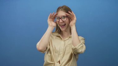 Mavi stüdyoda gülümseyen genç sarışın kadın elleri ve gözlükleriyle yüzünü örtüyor..