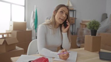Yeni bir evde notlar alırken telefonla konuşan bir kadın oturma odasındaki kutuları taşıyarak pencerelerden içeri doğal ışık giriyor..