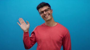 Gözlüklü ve kırmızı kazaklı gülümseyen adam mavi arka planda sıcak bir karşılama yapıyor..