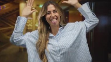 Zarif sarışın kadın parlak bir gülümseme ile kollarını açıyor ve bir otel lobisinde başını eğiyor pirinç sütunlar ve sıcak tepe ışıkları ile süslenmiş;.