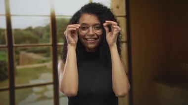 Siyah bluzlu, kıvırcık saçlı, gülümseyen genç bir kadın güneş ışığı altındaki stüdyo odasında gözlüğünü çıkartıyor..