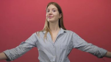 Çizgili gömlekli kadın kırmızı stüdyoda kameraya gülümserken başparmaklarını göğsüne gösteriyor..