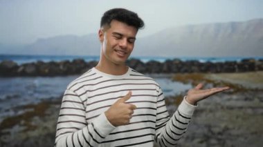 Sahilde duran genç adam başparmaklarını kaldırarak ve açık bir el göstererek arka planda dağlar ve bulutlu gökyüzü altında dalgalar olan çizgili tişört giyerek kıyıda duruyor.