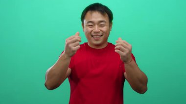 Kırmızı gömlekli genç adam yeşil arka plana karşı para hareketi yapıyor. İzole edilmiş bir stüdyoda başarıyı ve finansal refahı sembolize eden neşeli bir ifadeyle.