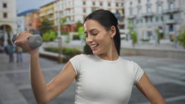 Şehir caddesinde halterle egzersiz yapan, arka planda canlı binalar olan, açık havada spor yapan, kendine güven ve neşe saçan bir kadın..